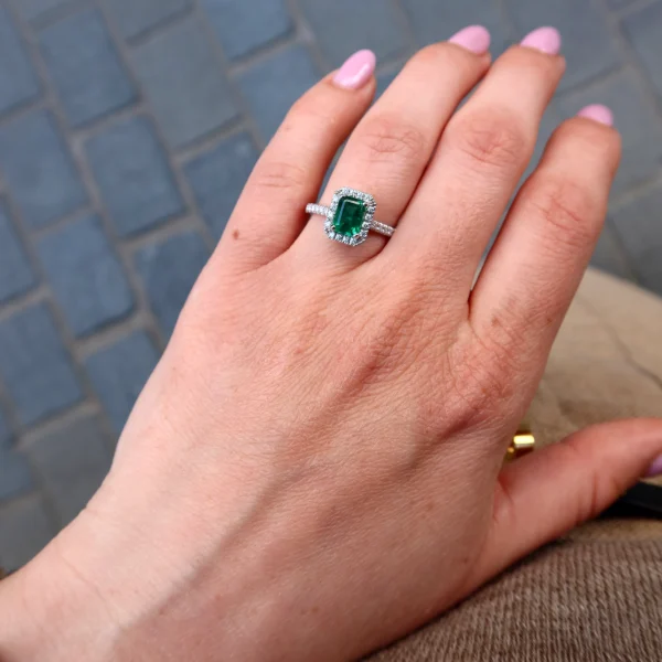 Hand shot of an emerald cut emerald halo ring in 18k white gold with pavé diamond band and rectangular green emerald centre stone.