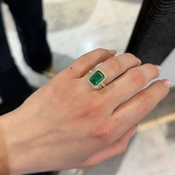 Hand wearing emerald cut emerald ring with diamond halo in yellow gold