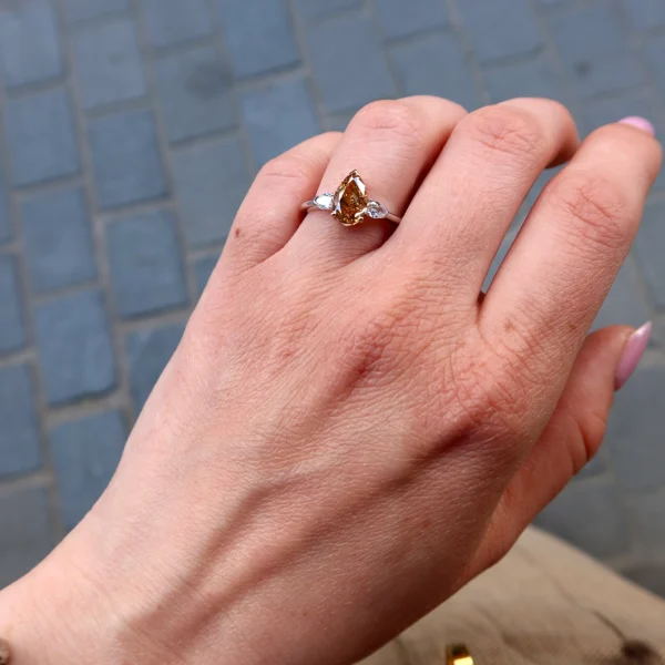 Bague diamant poire brun-orange portée à la main en or blanc et or rose 18 carats avec deux diamants latéraux taille poire.